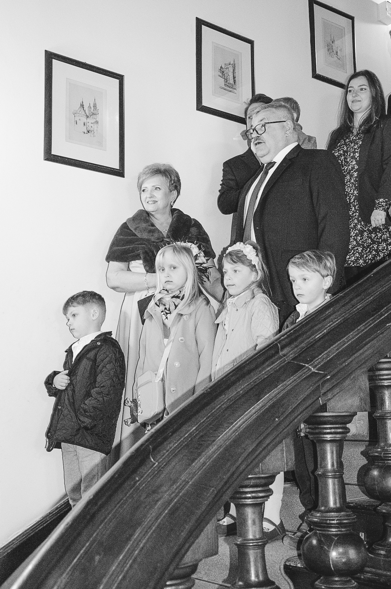 Czarno-białe zdjęcie grupy osób na schodach, w tym dzieci z opaskami na głowach, oraz dorosłych w formalnych strojach. W tle wiszą obrazy w ramkach.