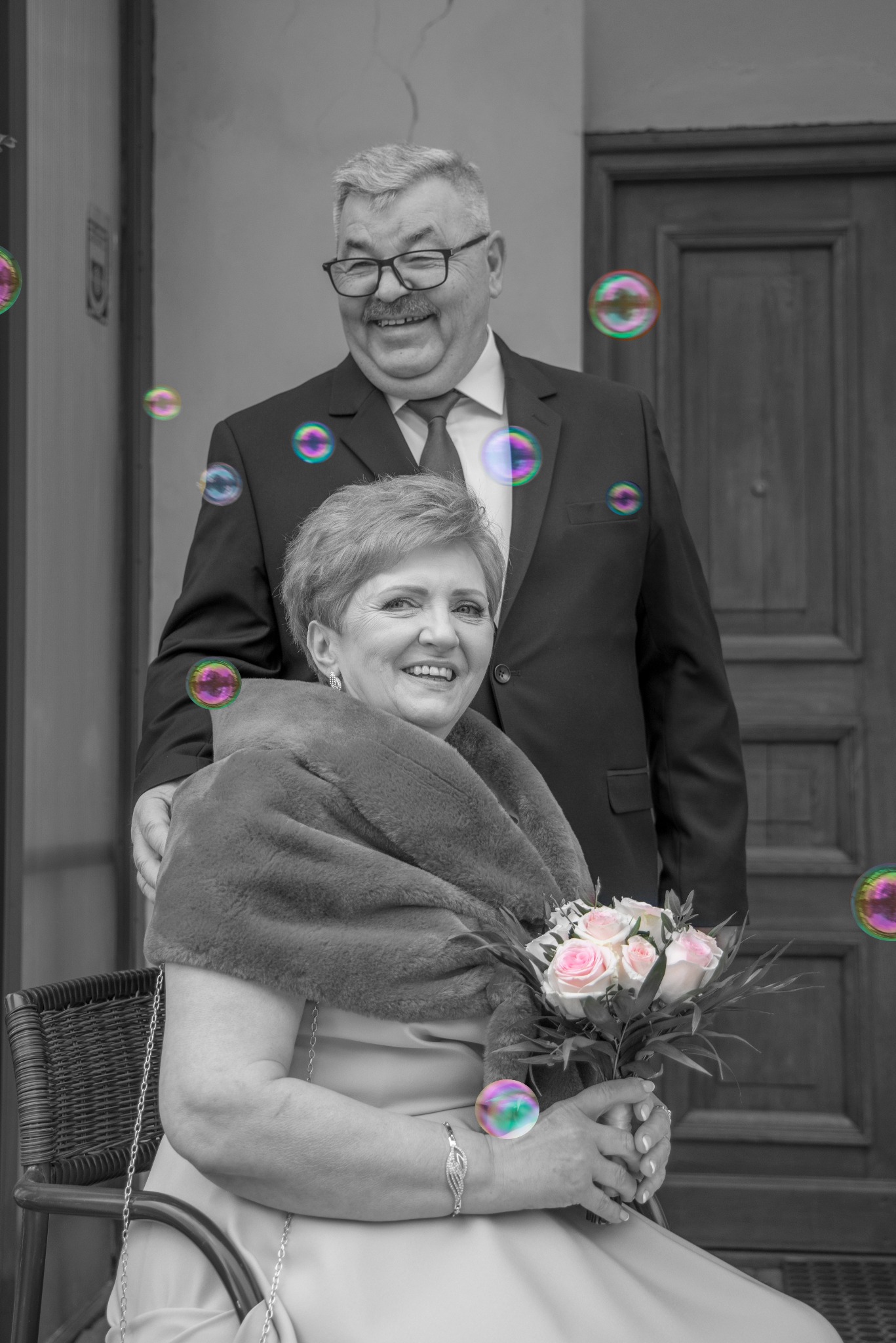 Czarno-białe zdjęcie uśmiechniętej pary w eleganckich strojach, otoczonej bańkami mydlanymi. Kobieta siedzi z bukietem różowych róż, mężczyzna stoi za nią, obejmując ją.