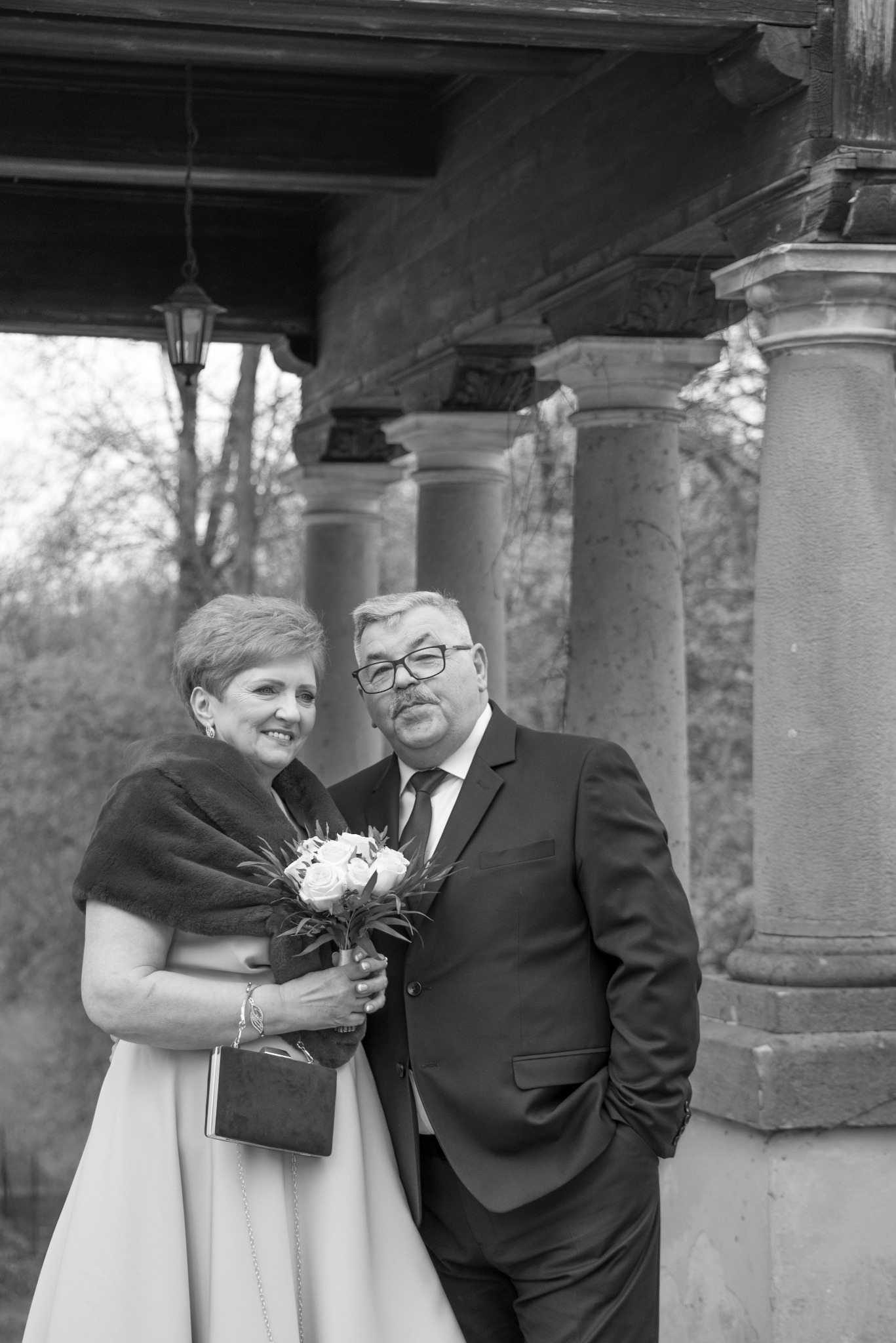 Czarno-białe zdjęcie eleganckiej pary w średnim wieku, pozującej z bukietem róż pod kolumnadą. Kobieta trzyma małą torebkę. Uśmiechnięci, w tle drzewa.