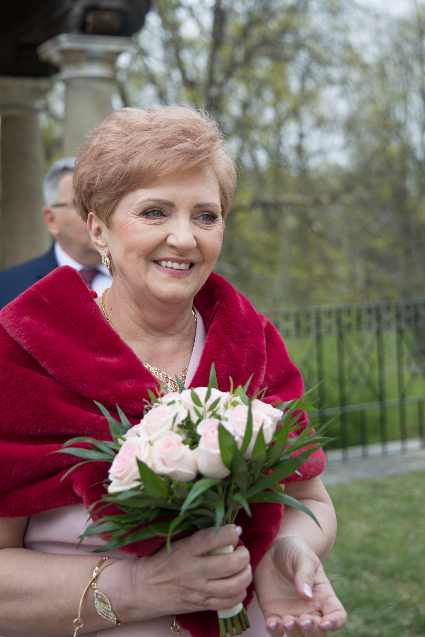 Uśmiechnięta kobieta z krótkimi włosami, w czerwonym szalu, trzyma bukiet różowych róż. W tle widoczny mężczyzna i zieleń.