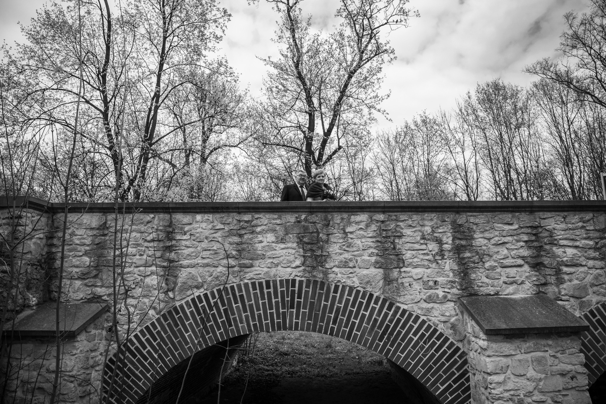 Czarno-białe zdjęcie pary na kamiennym moście z ceglanym łukiem, otoczonego drzewami bez liści, pod pochmurnym niebem. Ujęcie z dołu, podkreślające architekturę.