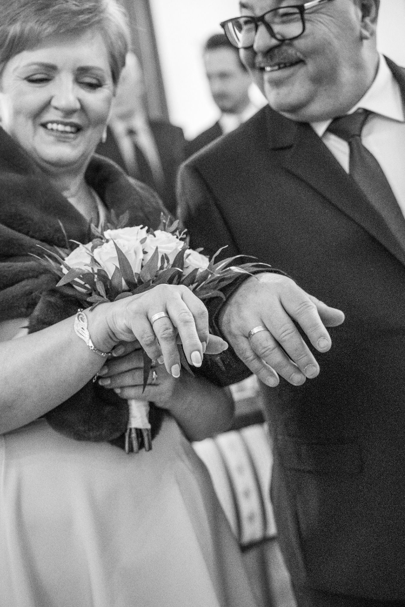 Czarno-białe zbliżenie dłoni pary młodej z obrączkami, bukiet ślubny w dłoniach kobiety, uśmiechnięte twarze w tle, uroczysty nastrój.
