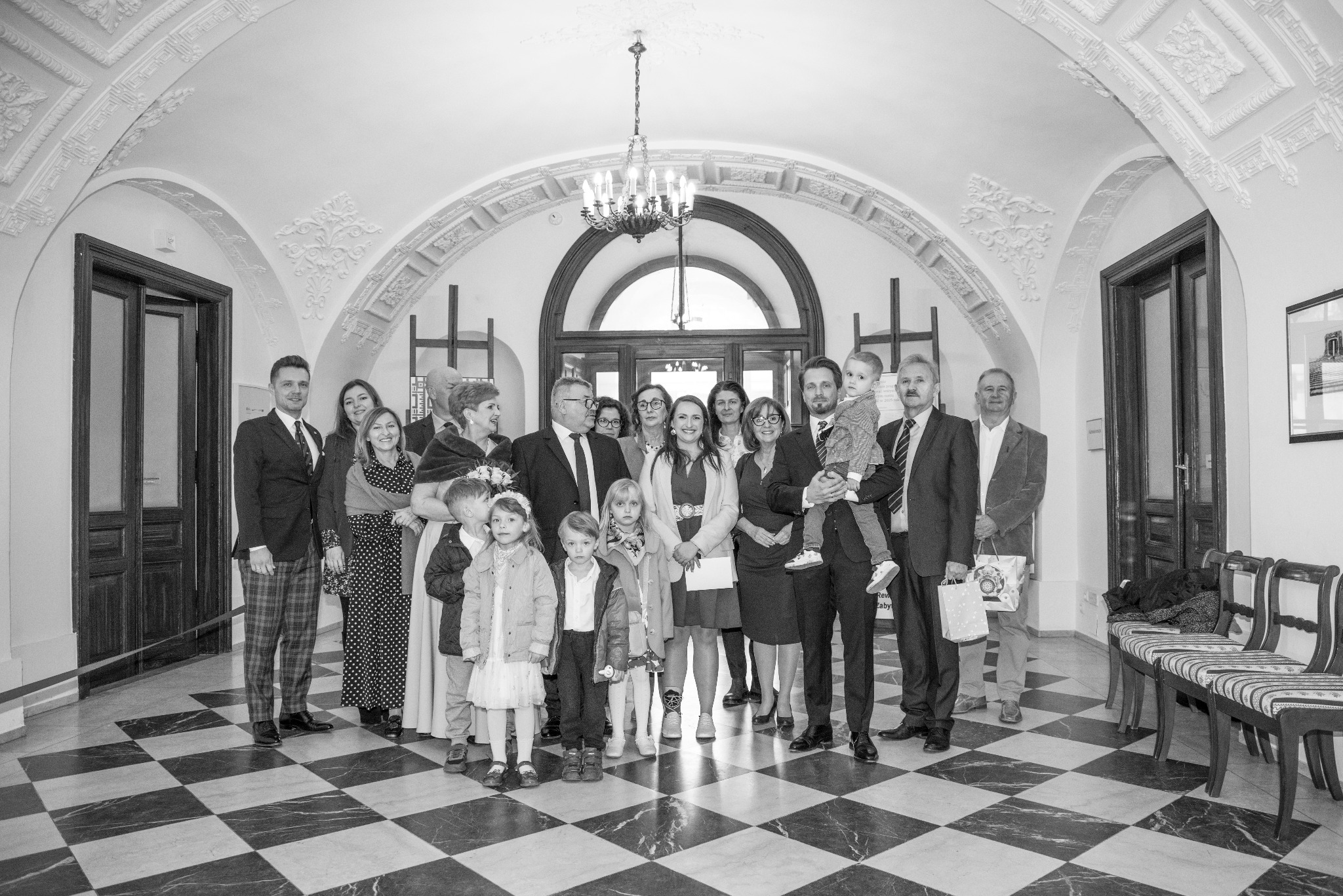 Czarno-białe zdjęcie grupowe w eleganckim wnętrzu z marmurową podłogą w szachownicę, wysokimi sufitami i ozdobnymi łukami. W centrum uśmiechnięta kobieta.