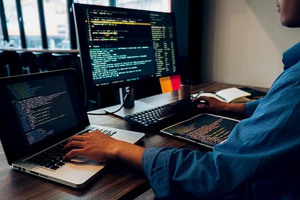 Programista pracuje na dwóch monitorach, pisząc kod. Na biurku laptop, notatnik i tablet graficzny. Widoczne karteczki samoprzylepne i kubek.