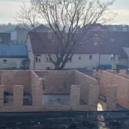 Wczesny etap budowy domu z czerwonej cegły, widoczne fundamenty i pierwsze ściany, w tle zabudowa jednorodzinna i drzewa w słoneczny dzień. Praca w toku.