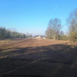 Przygotowany grunt pod zasiew trawy, widoczne ślady opon, w tle dom i drzewa bez liści, błękitne niebo. Jesienne słońce oświetla pole.