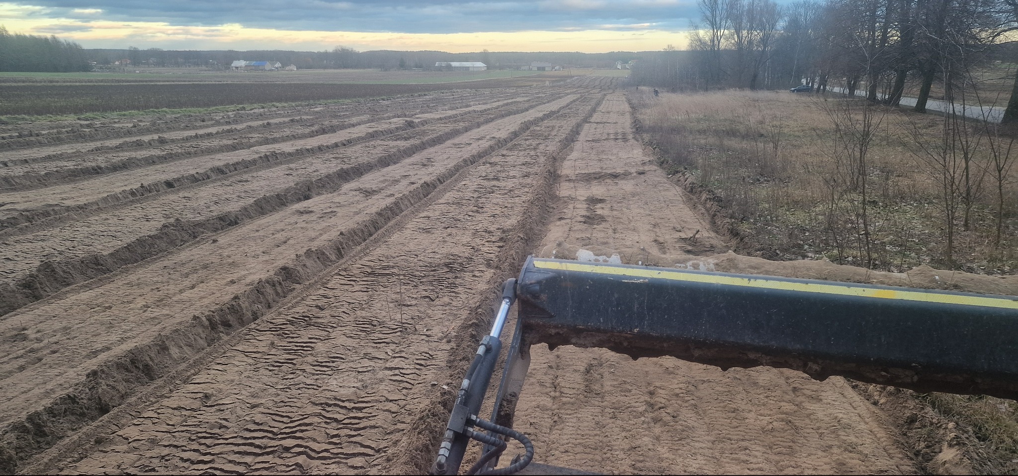 Niwelacja terenu: Równoległe bruzdy w ziemi, wykonane ciężkim sprzętem, przygotowane pod uprawę. Widok z perspektywy operatora maszyny.