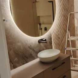 Nowoczesna łazienka z okrągłym lustrem z podświetleniem LED, umywalką nablatową i mozaiką w jodełkę na ścianie. Widoczna drabina wskazuje na trwające prace wykończeniowe.