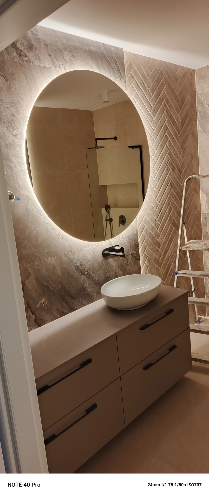 Nowoczesna łazienka z okrągłym lustrem z podświetleniem LED, umywalką nablatową i mozaiką w jodełkę na ścianie. Widoczna drabina wskazuje na trwające prace wykończeniowe.