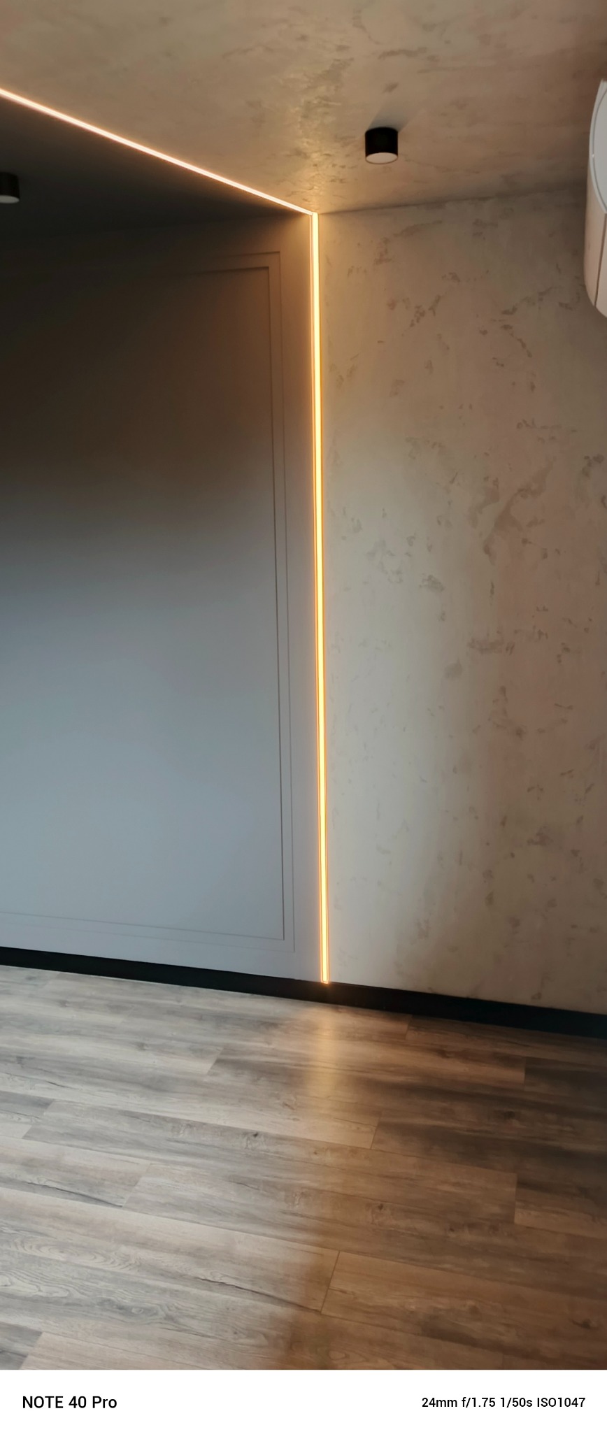 Minimalistyczne wnętrze po remoncie: szara ściana z ukrytą listwą LED, drewnopodobna podłoga i czarny plafon. Nowoczesne wykończenie.