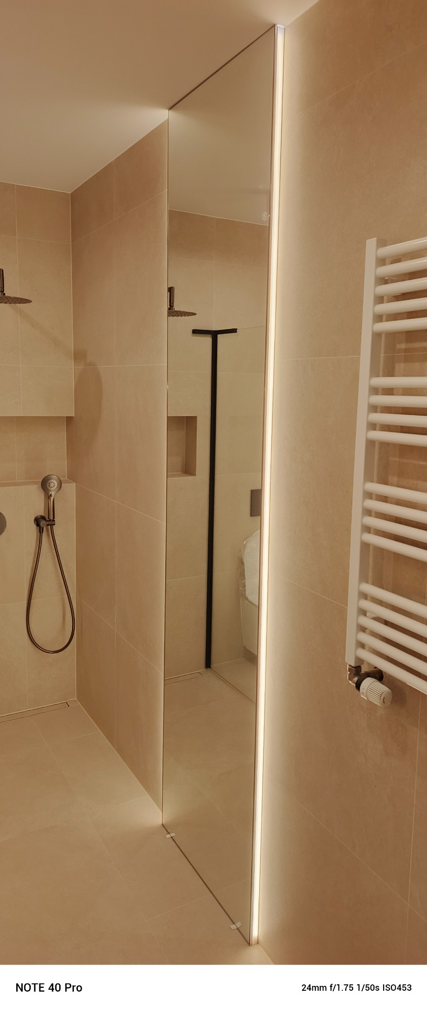 Nowoczesna łazienka z kabiną typu walk-in, beżowe płytki, oświetlenie LED wzdłuż szyby kabiny, grzejnik drabinkowy. Minimalistyczny design.