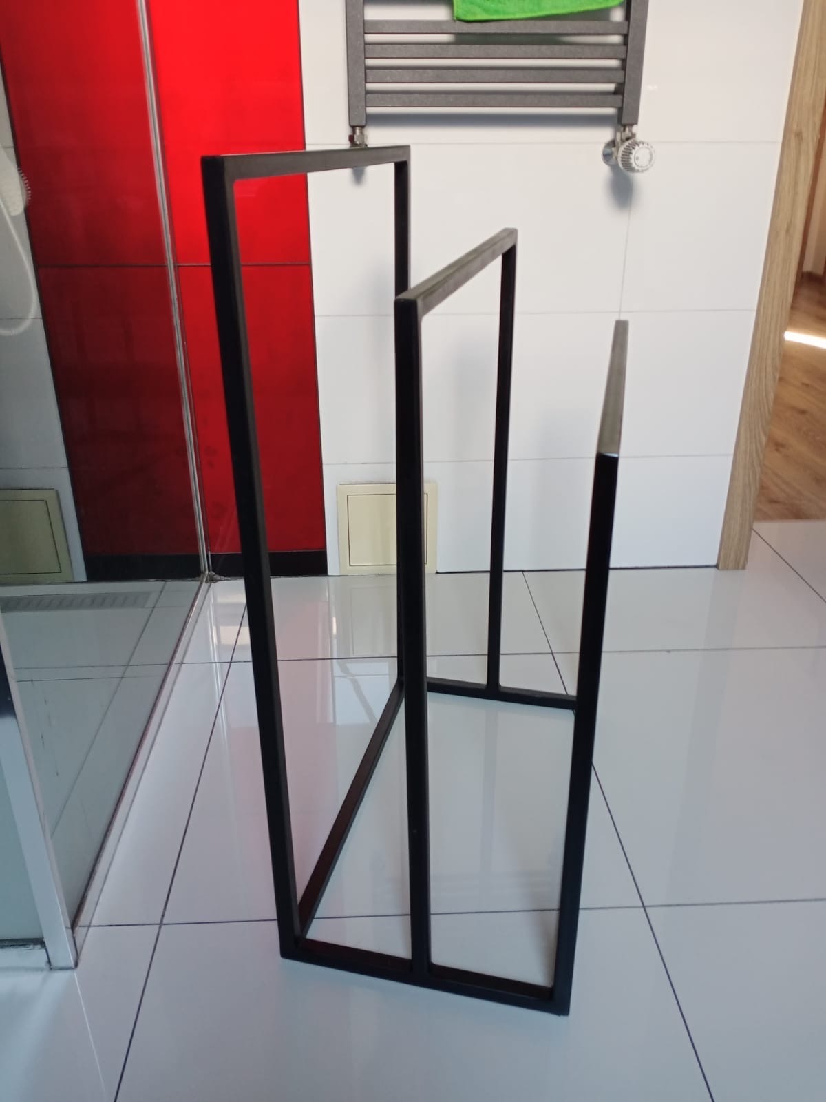 Czarny, metalowy stojak na ręczniki w łazience z białą glazurą i grzejnikiem. Minimalistyczny design, spawana konstrukcja, nowoczesne wykończenie.