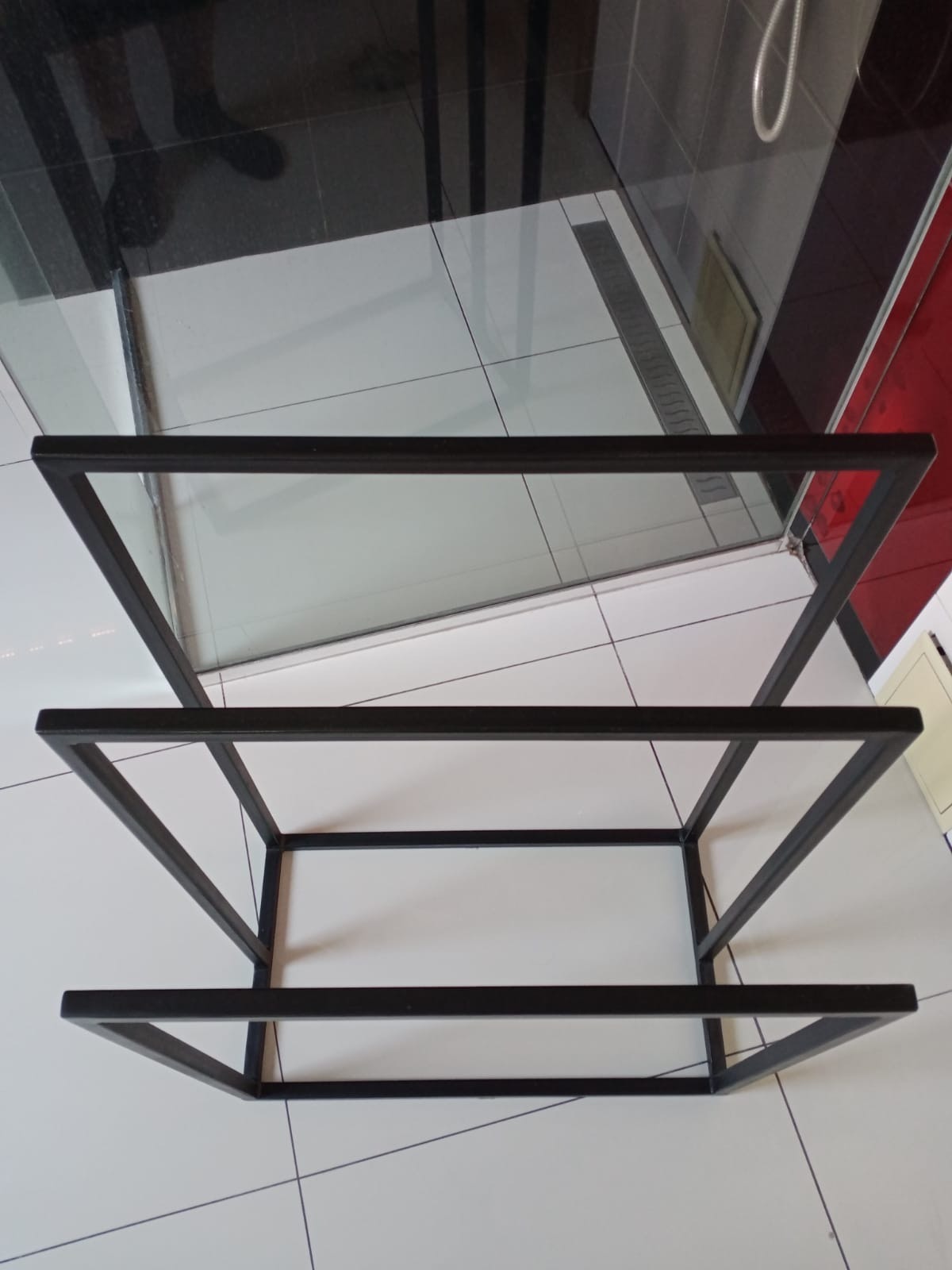 Czarna, metalowa konstrukcja regału z trzema półkami na tle białych i czarnych płytek w łazience. Widoczne mocne spawy na rogach.