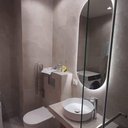 Remonty Pod Klucz - Ukończona łazienka z beżowymi płytkami, owalnym lustrem z podświetleniem LED, umywalką nablatową i kabiną prysznicową ze szkła. W tle pralka.