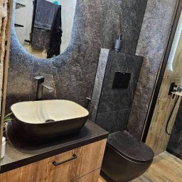 Remonty Pod Klucz - Nowoczesna łazienka z umywalką nablatową, toaletą w kolorze antracytu i lustrem z podświetleniem LED. Na ścianach szare płytki imitujące marmur.