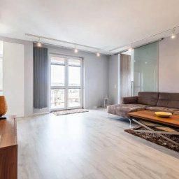 Remonty Pod Klucz - Jasne, przestronne wnętrze salonu z telewizorem na ścianie, drewnianą komodą, kanapą i stolikiem kawowym. Duże okno balkonowe z grzejnikiem. Nowoczesne oświetlenie sufitowe.