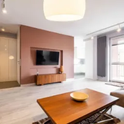 Jasne wnętrze salonu z drewnianymi meblami, ścianą w odcieniach terakoty i dużym telewizorem. Widoczne okno balkonowe z grzejnikiem i nowoczesne oświetlenie sufitowe.