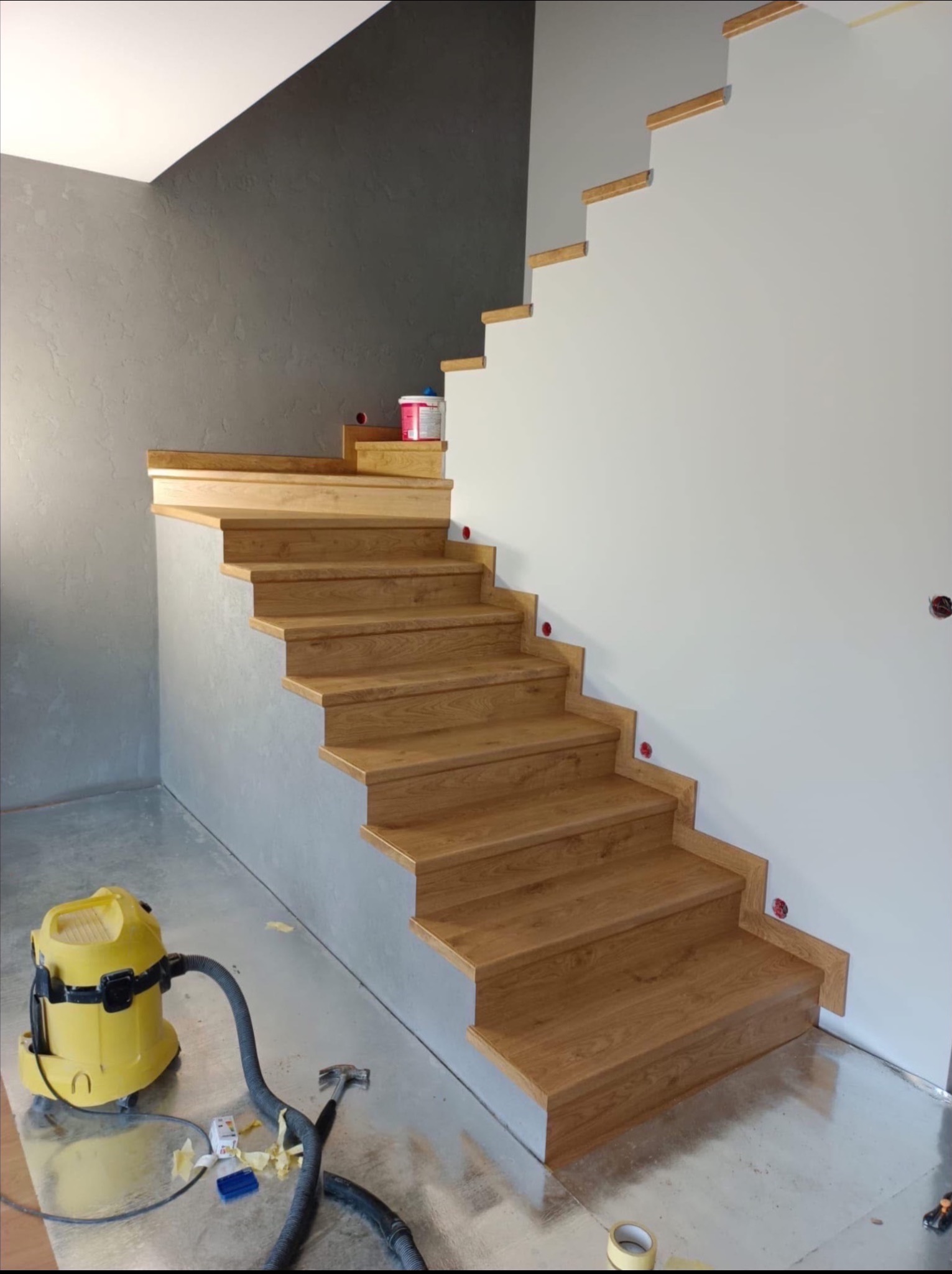 Drewniane schody z oświetleniem stopni, widok z dołu. Na podłodze folia zabezpieczająca i odkurzacz przemysłowy. Ściana szara, teksturowana. Detal wykończenia.