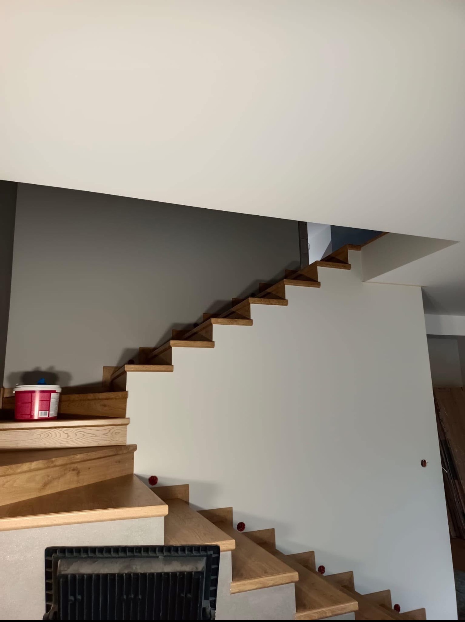 Drewniane schody z białymi podstopnicami w trakcie montażu, widoczne czerwone zaślepki. Ściana pomalowana na szaro, wiadro z farbą na pierwszym stopniu.