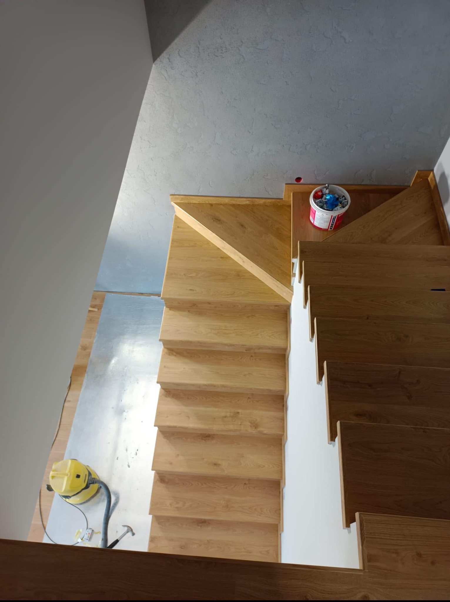 Drewniane schody po cyklinowaniu, widok z góry. Odkurzacz i wiadro z farbą obok. Jasne drewno, szare ściany i białe podstopnice. Etap wykończenia.