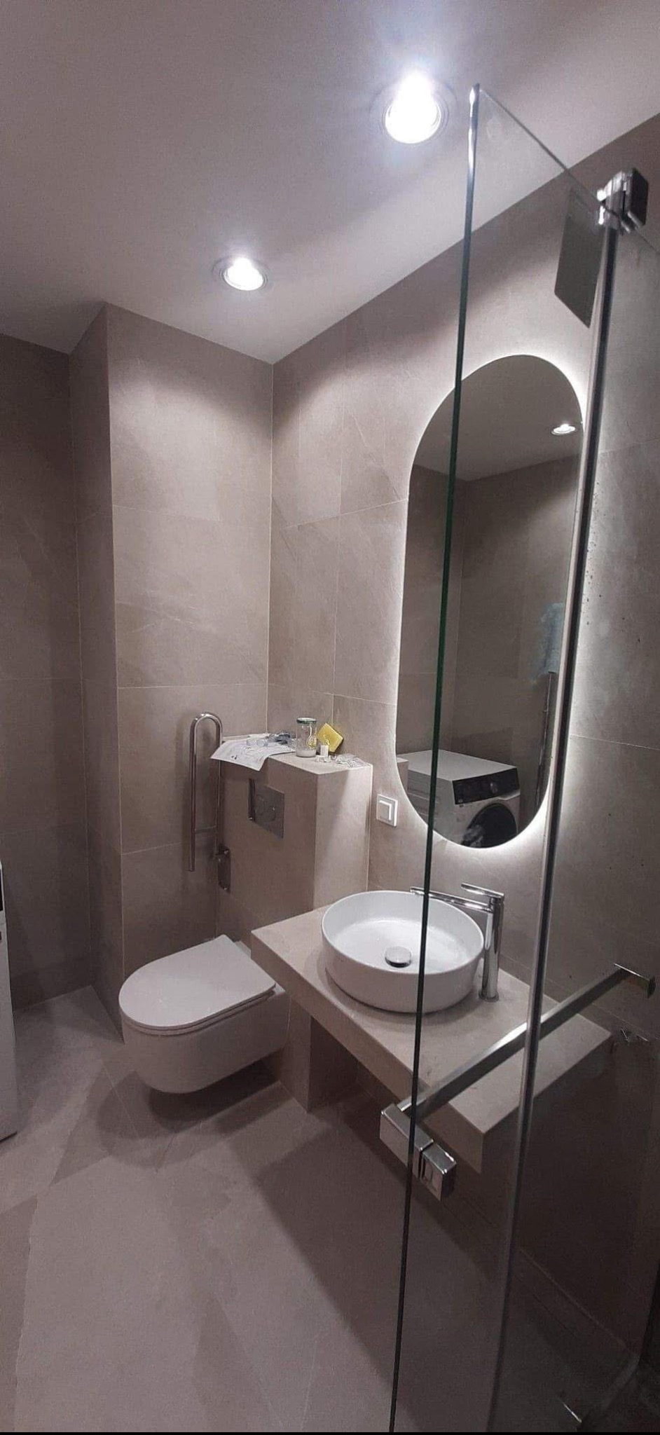 Ukończona łazienka z beżowymi płytkami, owalnym lustrem z podświetleniem LED, umywalką nablatową i kabiną prysznicową ze szkła. W tle pralka.