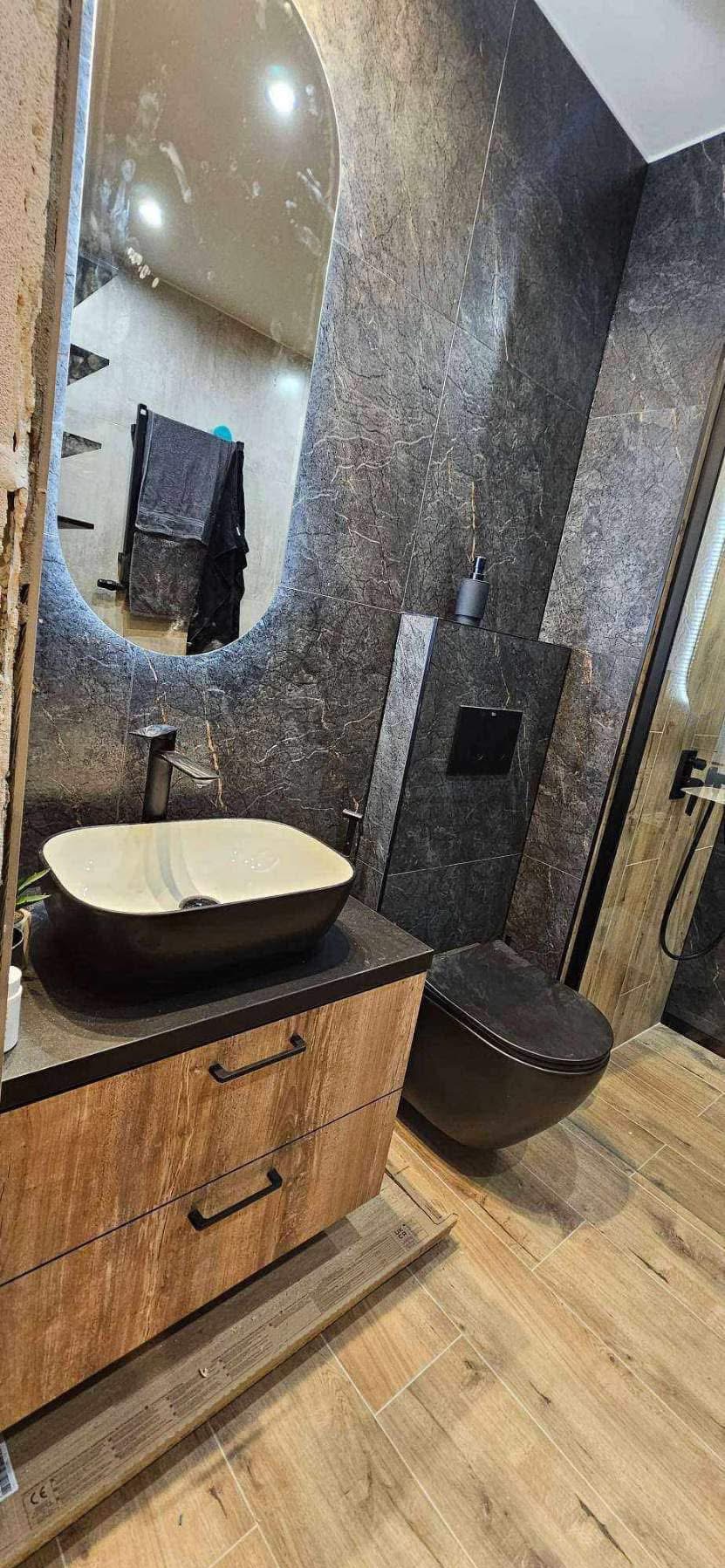 Nowoczesna łazienka z umywalką nablatową, toaletą w kolorze antracytu i lustrem z podświetleniem LED. Na ścianach szare płytki imitujące marmur.