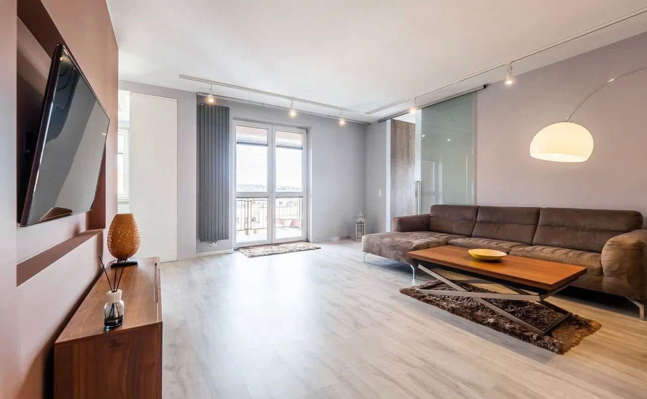 Jasne, przestronne wnętrze salonu z telewizorem na ścianie, drewnianą komodą, kanapą i stolikiem kawowym. Duże okno balkonowe z grzejnikiem. Nowoczesne oświetlenie sufitowe.