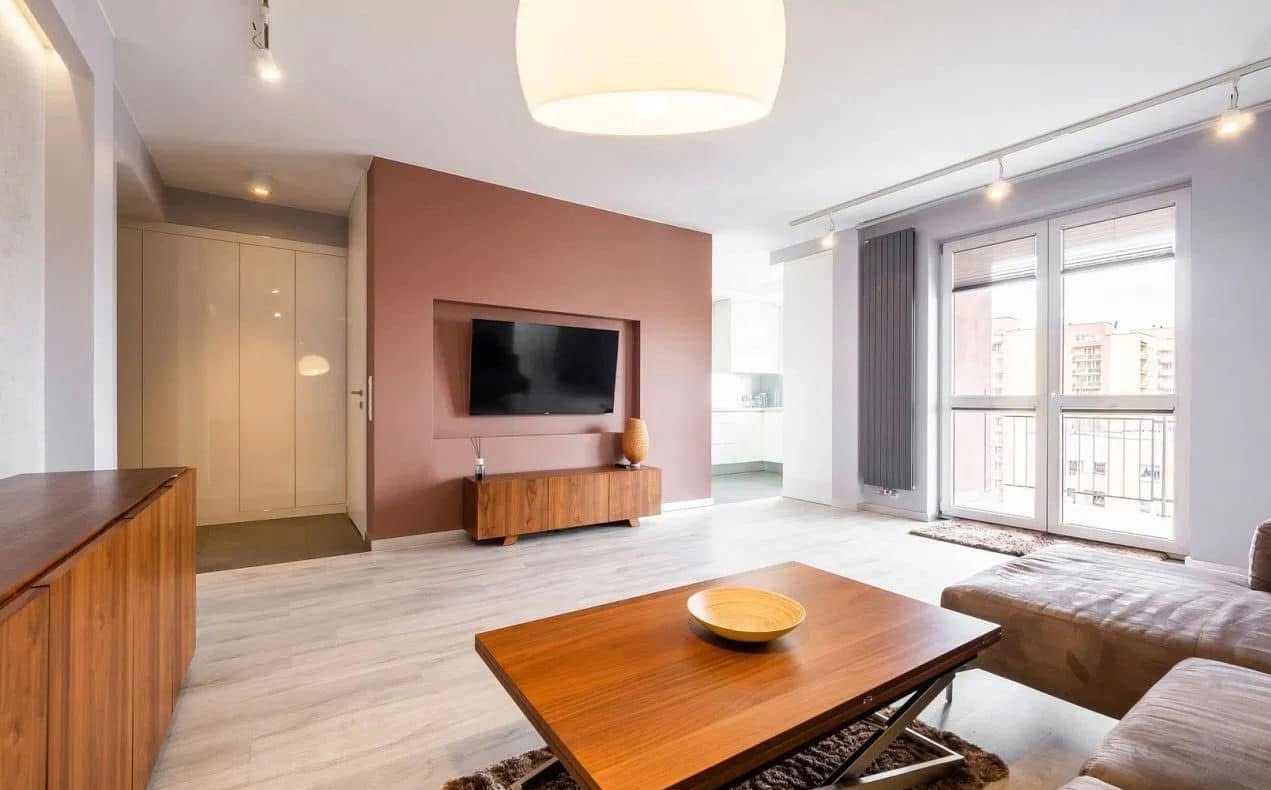 Jasne wnętrze salonu z drewnianymi meblami, ścianą w odcieniach terakoty i dużym telewizorem. Widoczne okno balkonowe z grzejnikiem i nowoczesne oświetlenie sufitowe.