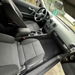 Wnętrze czarnego samochodu osobowego marki Audi z szarą tapicerką tekstylną. Widoczne fotele, deska rozdzielcza i kierownica. Perspektywa z zewnątrz auta.