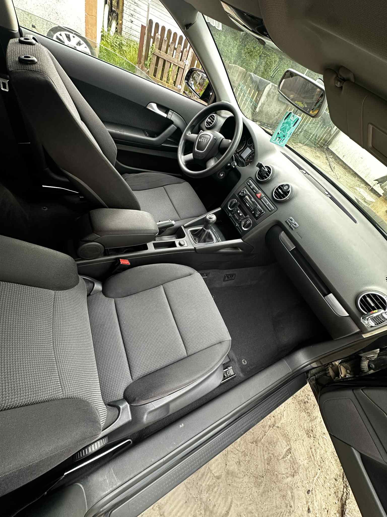 Wnętrze czarnego samochodu osobowego marki Audi z szarą tapicerką tekstylną. Widoczne fotele, deska rozdzielcza i kierownica. Perspektywa z zewnątrz auta.