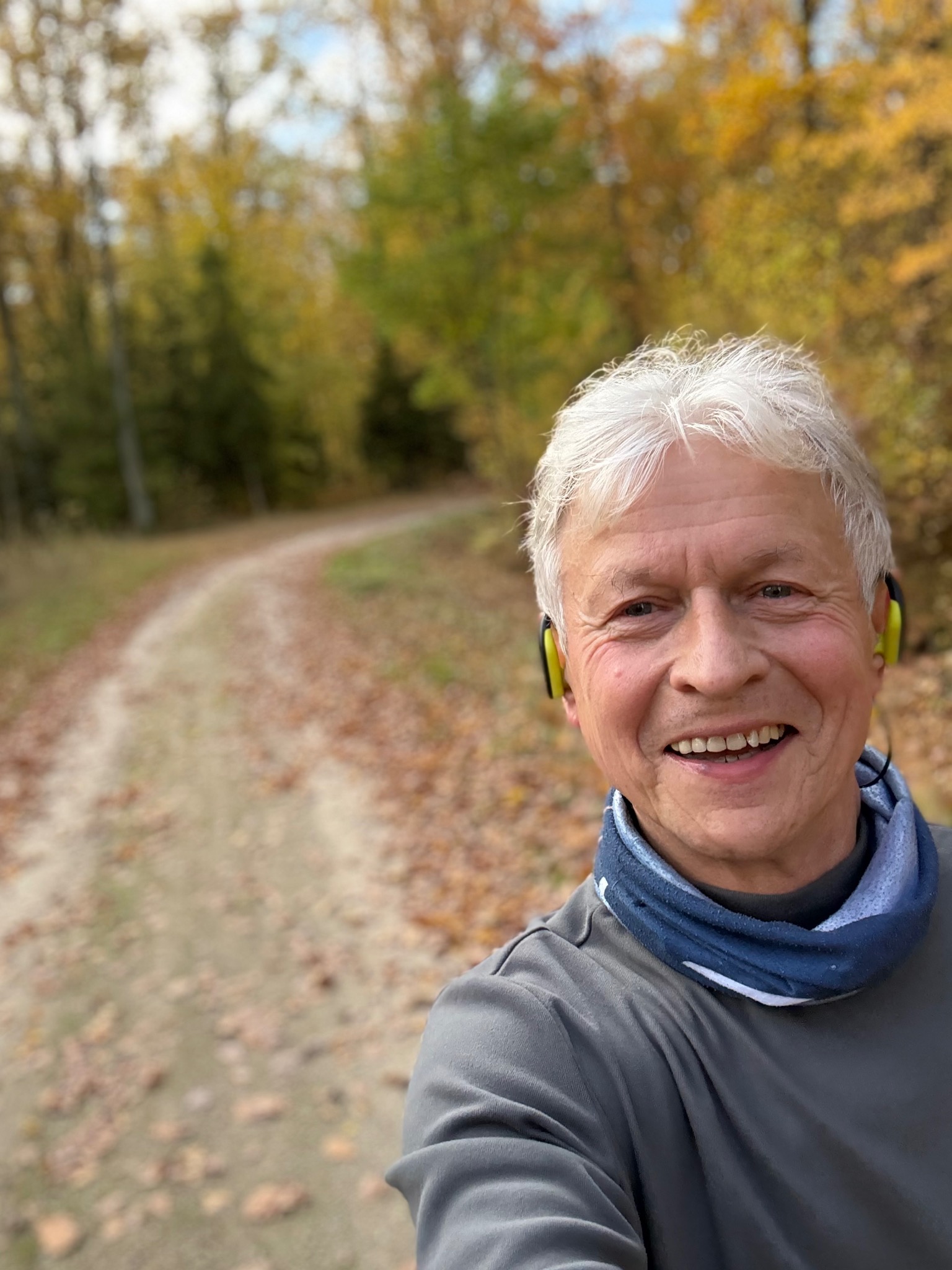 Uśmiechnięty mężczyzna z siwymi włosami w słuchawkach, robiący selfie na leśnej ścieżce w jesiennej scenerii. Widoczne opadłe liście i drzewa w tle.