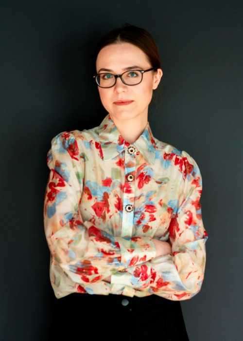 Portret kobiety w okularach, adwokat ze Śremu, w bluzce w kwiaty, z założonymi rękami na tle ciemnej ściany. Profesjonalny wizerunek prawnika.