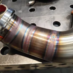 Precyzyjnie spawany, nierdzewny element rurowy z widocznymi tęczowymi przebarwieniami spoin, leżący na perforowanym stole spawalniczym. Bliski plan, ostre światło.