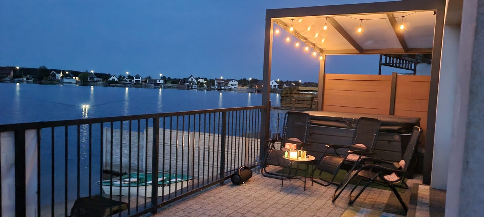 Wieczorny relaks na balkonie z metalową barierką, oświetlonym pergolą i widokiem na jezioro. W tle domy odbijające się w wodzie.
