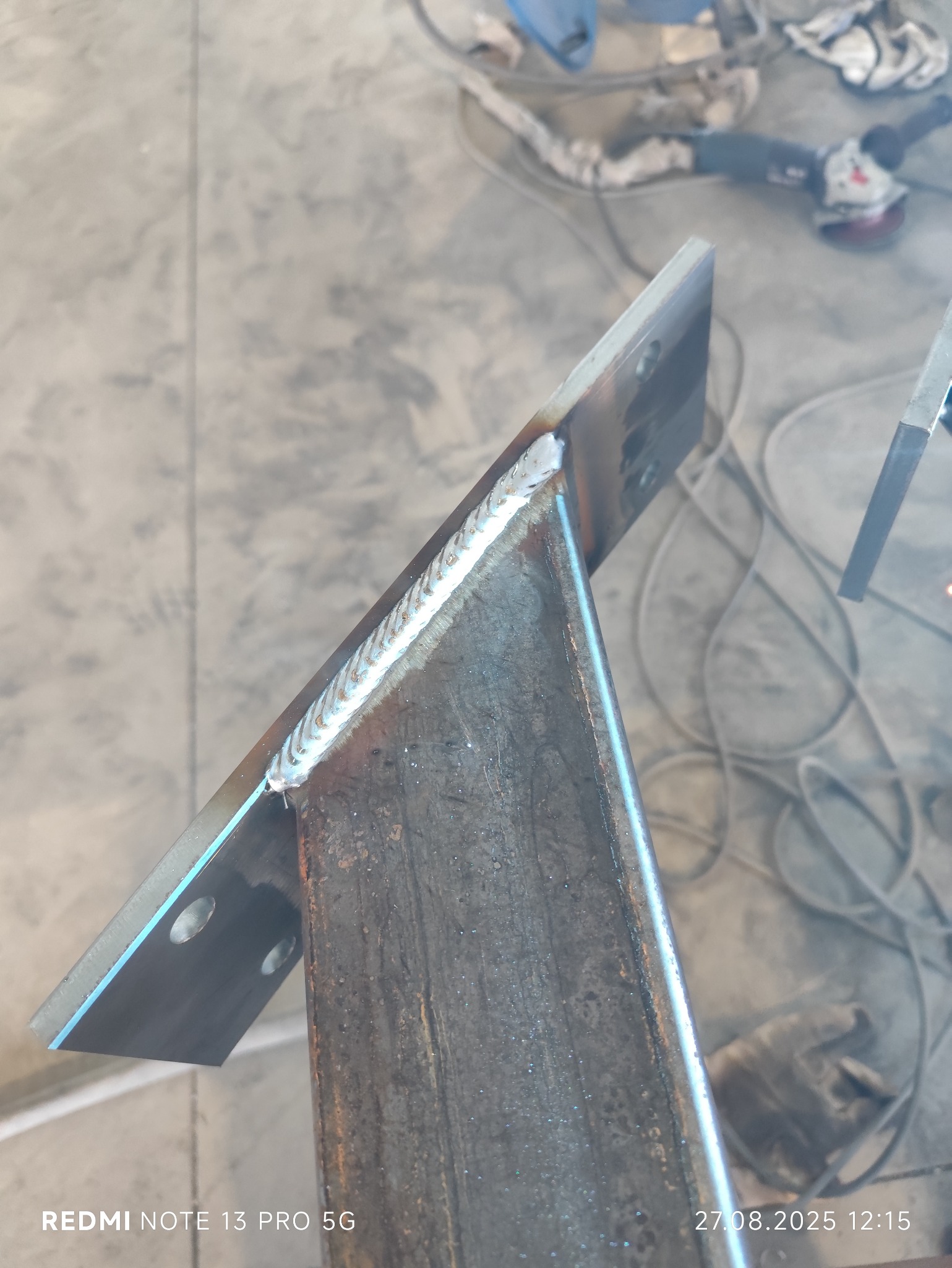 Spoina na metalowym elemencie konstrukcyjnym, widoczne ślady spawania. W tle szlifierka kątowa i przewody.