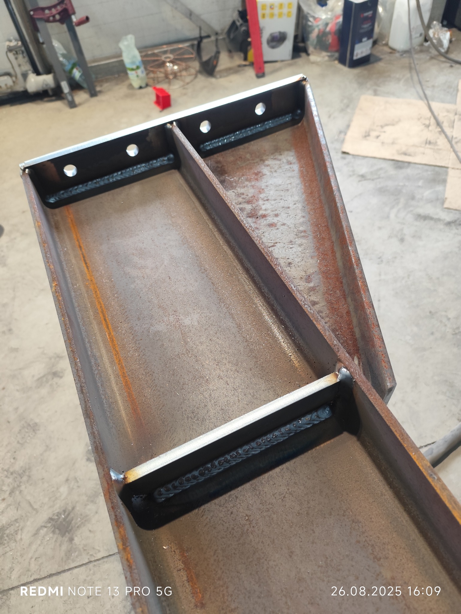 Metalowa konstrukcja z widocznymi spawami i otworami, wykonana z surowej stali. Element konstrukcyjny w warsztacie, prawdopodobnie w trakcie tworzenia.