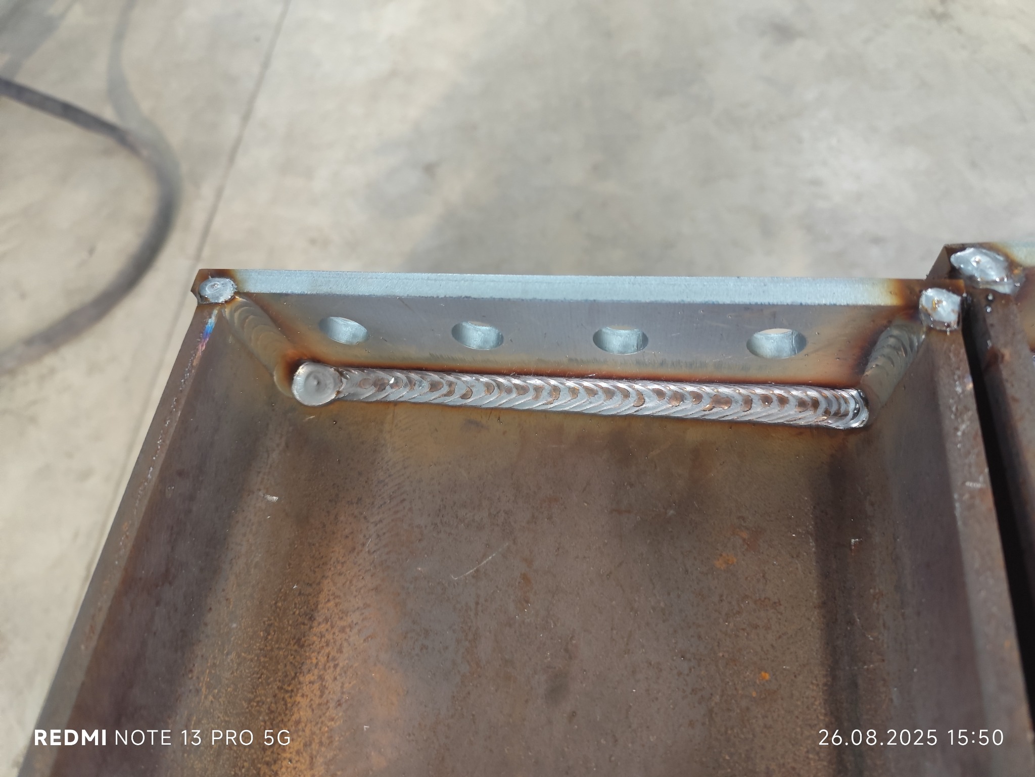 Spoina łącząca metalowe elementy, widoczne otwory w górnej płycie. Charakterystyczny wzór spawu. Metal o surowym wykończeniu, widoczne ślady obróbki.