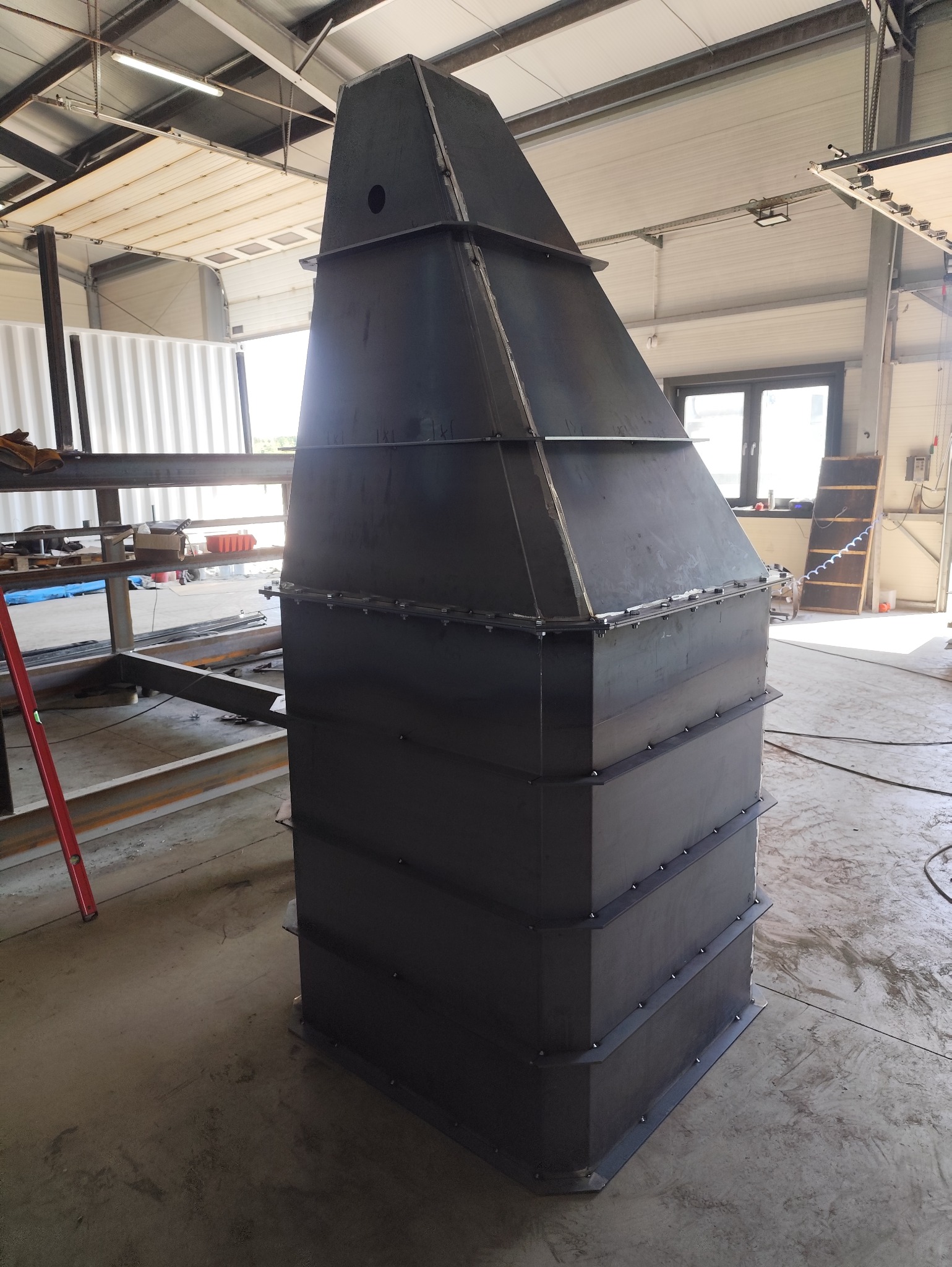 Konstrukcja stalowa w warsztacie. Metalowa, spawana konstrukcja o trapezoidalnym kształcie. Widoczne spawy i elementy złączne. Hala produkcyjna w tle.