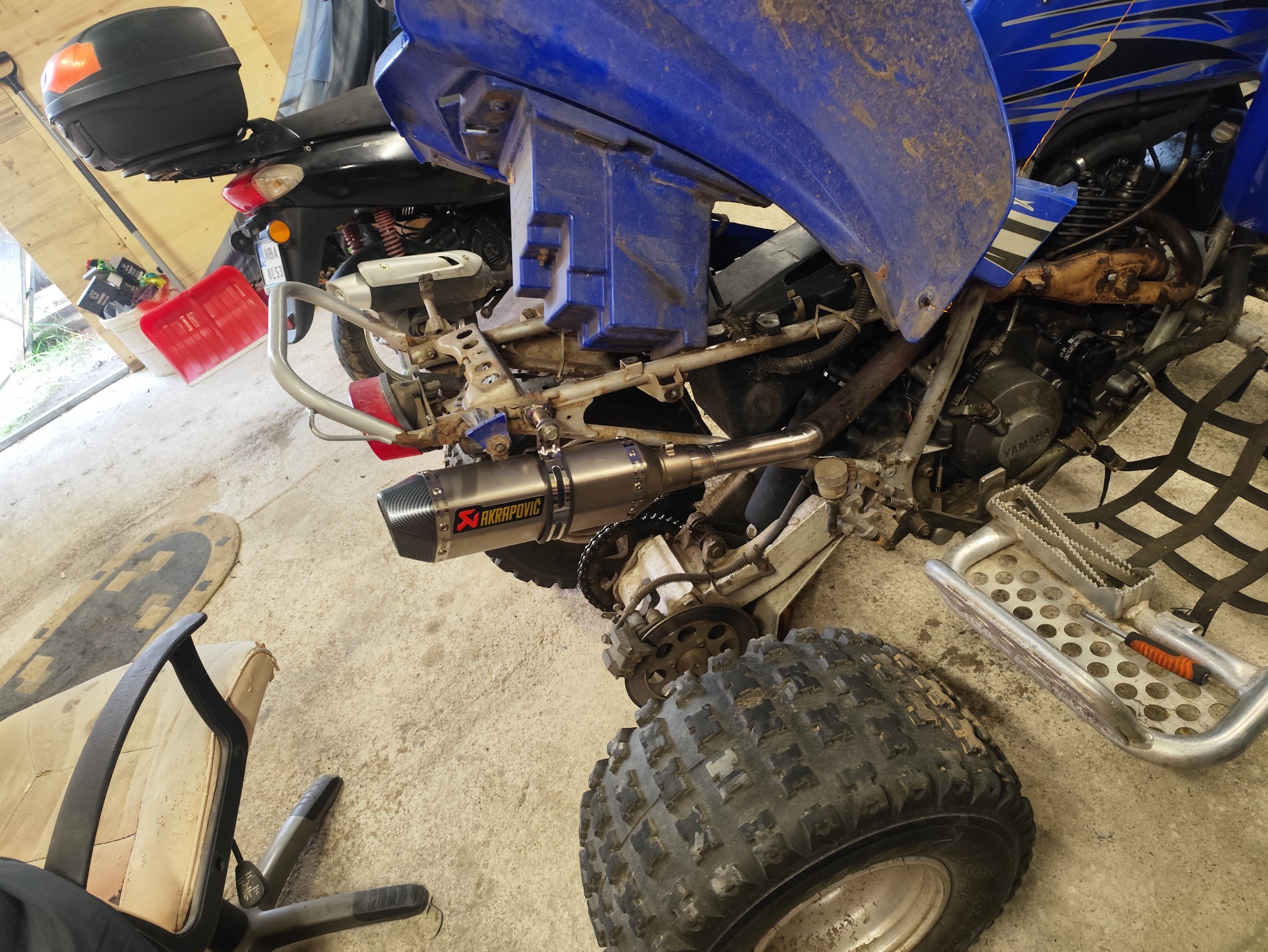 Nietypowe ujęcie quada Yamaha z zamontowanym wydechem Akrapovic, widoczne detale konstrukcyjne i części silnika. Zdjęcie z garażu.