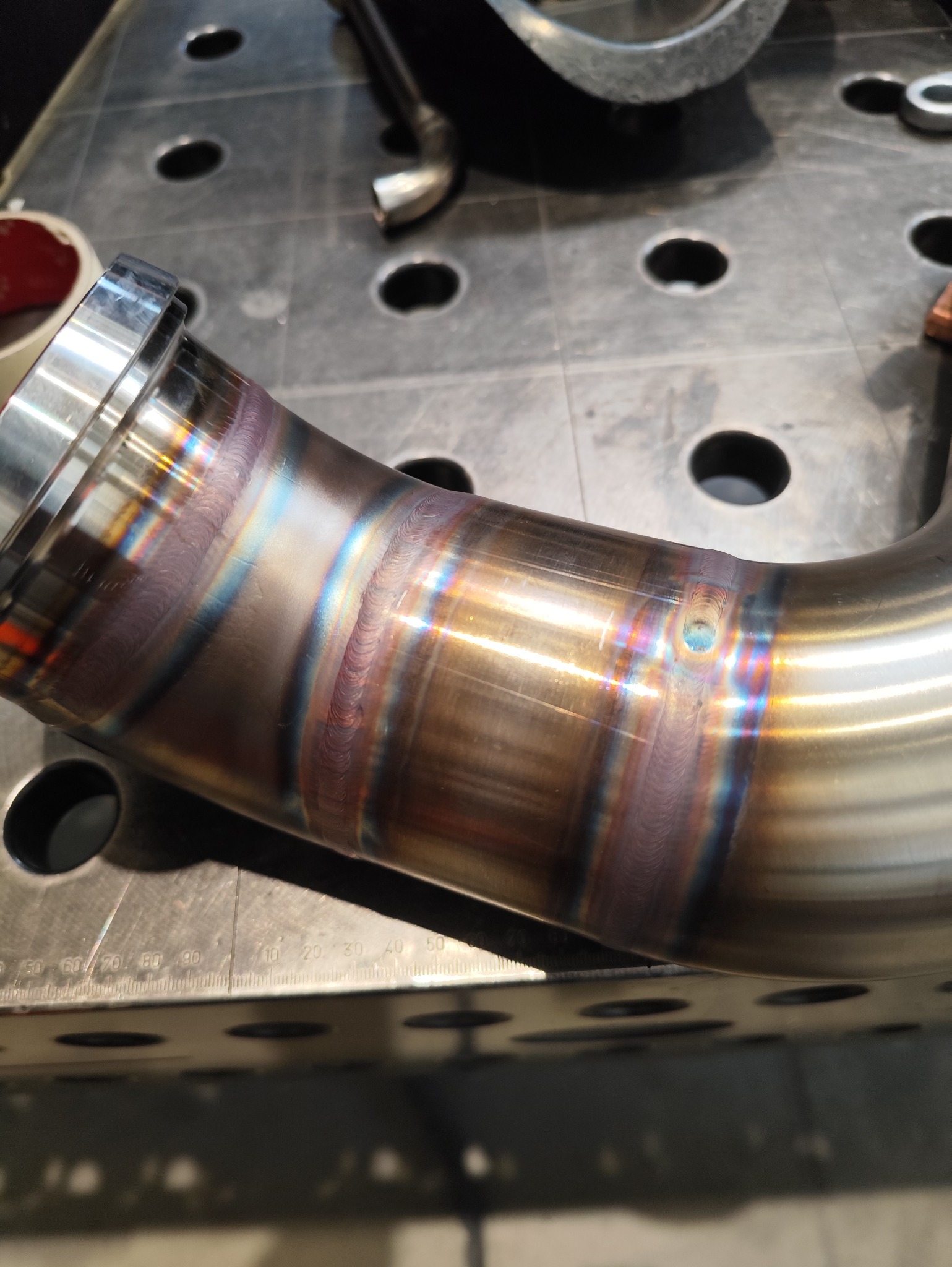 Precyzyjnie spawany, nierdzewny element rurowy z widocznymi tęczowymi przebarwieniami spoin, leżący na perforowanym stole spawalniczym. Bliski plan, ostre światło.