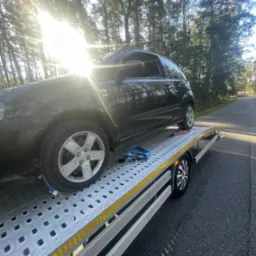 Laweta z czarnym samochodem osobowym na metalowej platformie, zabezpieczonym pasami, na tle leśnej drogi w słoneczny dzień.
