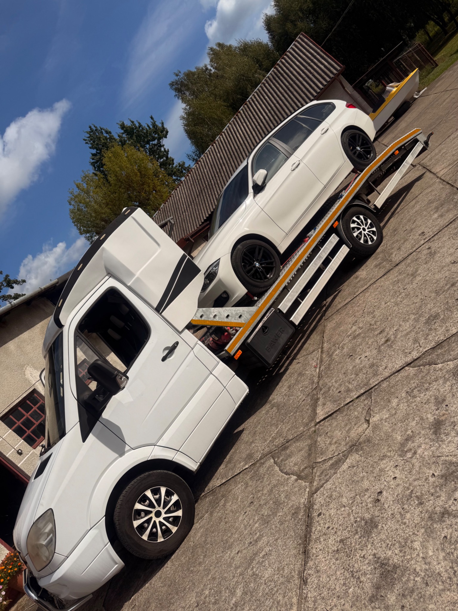 Biała laweta z białym BMW na platformie, na tle budynku w Ostrołęce. Auto z czarnymi felgami. Usługa transportu samochodów.