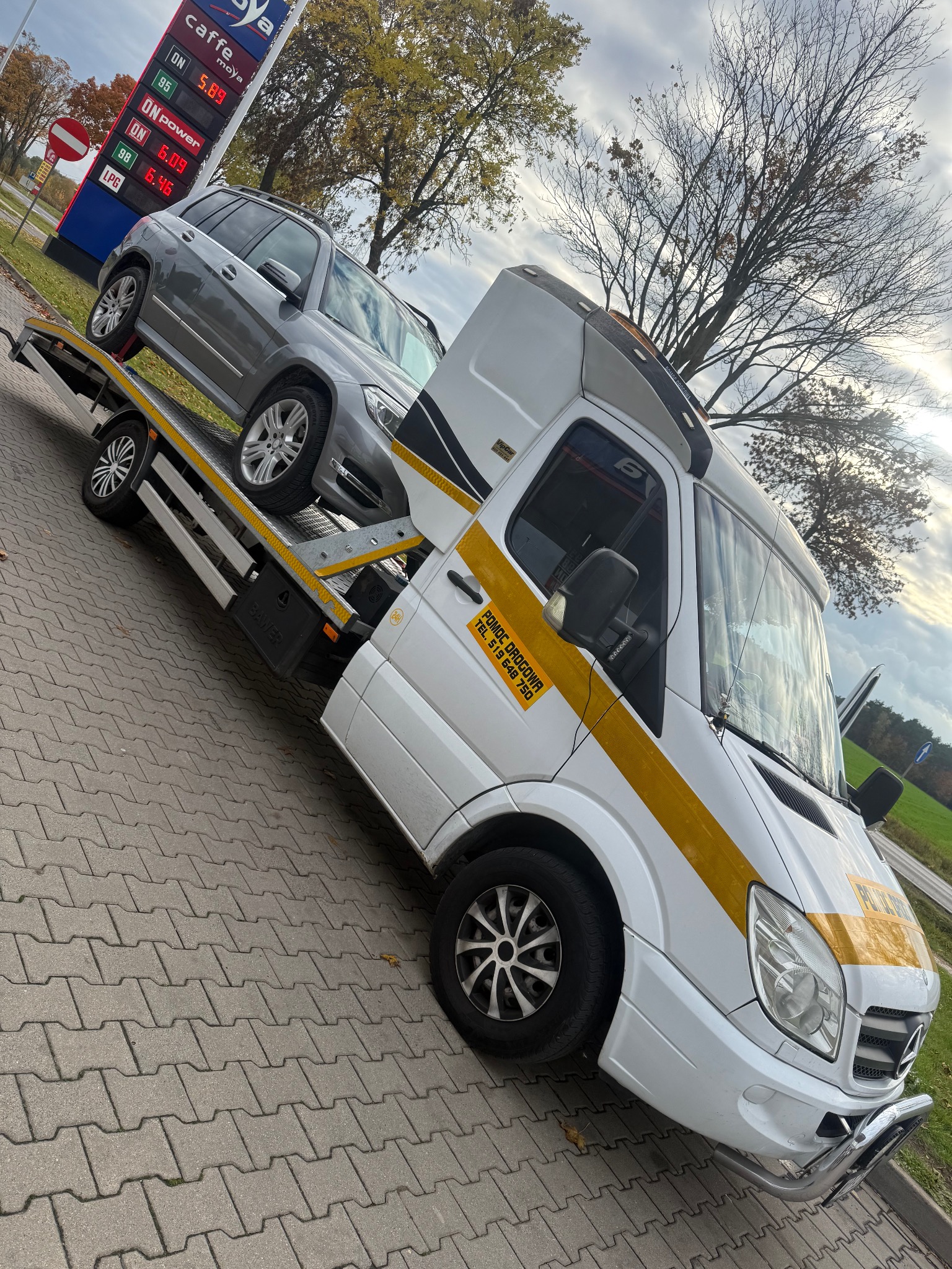 Biała laweta Mercedes z żółtymi pasami i napisem 'Pomoc Drogowa' przewozi szare auto osobowe na tle stacji paliw Moya i drzew.