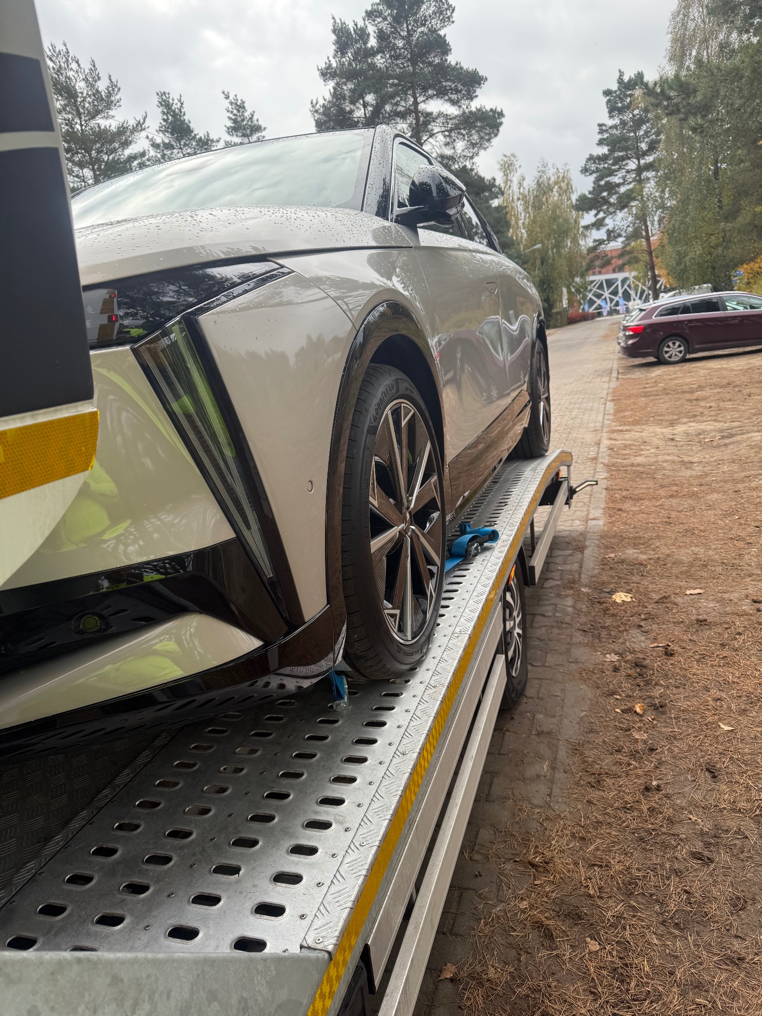 Nowoczesny, srebrny samochód zabezpieczony pasami na metalowej platformie lawety, widok z boku, jesienne tło z drzewami i chodnikiem.