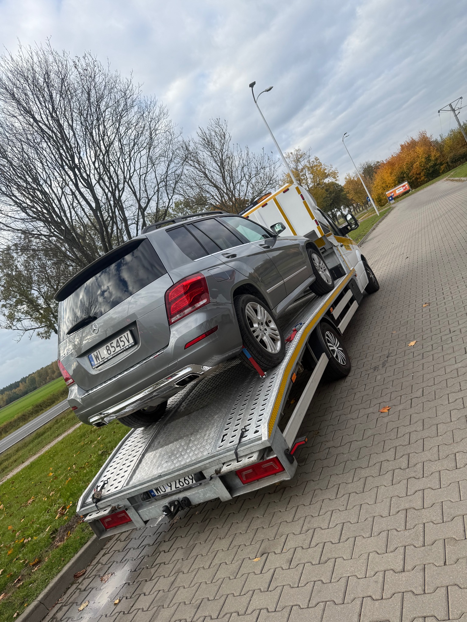 Szary Mercedes GLK na lawecie, widok z tyłu. Tablice rejestracyjne WL 8545V i WU 92669. Jesienne tło z drzewami i chodnikiem z kostki.