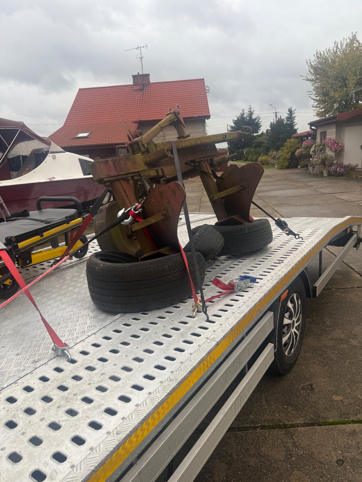 Laweta z ładunkiem: stary pług rolniczy zabezpieczony pasami transportowymi. W tle dom z czerwoną dachówką i fragment łodzi.