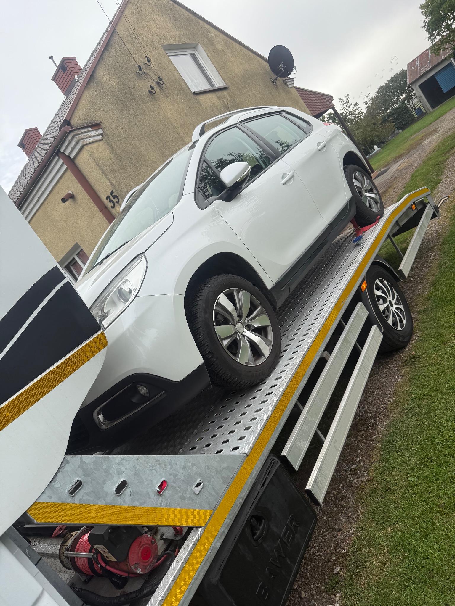 Biały samochód na lawecie przed domem z numerem 35. Widoczny hak holowniczy i trawa. Laweta z wyciągarką, gotowa do transportu pojazdu.