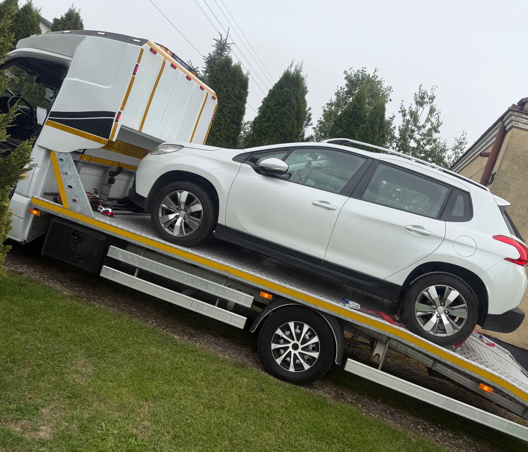 Biały Peugeot 2008 na platformie lawety z otwartą kabiną, zabezpieczony pasami transportowymi, na tle zieleni i pochmurnego nieba. Usługa lawetowania.