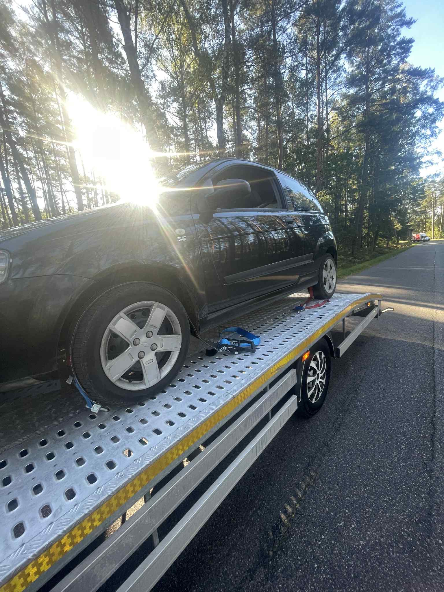 Laweta z czarnym samochodem osobowym na metalowej platformie, zabezpieczonym pasami, na tle leśnej drogi w słoneczny dzień.