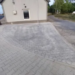 Szara kostka brukowa ułożona przed budynkiem z widocznymi paletami i fragmentem ulicy. Krawężnik oddziela chodnik od jezdni.